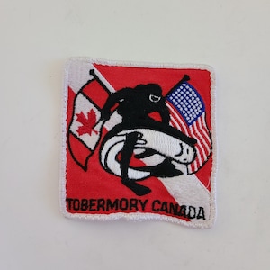 May include: A red and white embroidered patch with a diver holding a Canadian and American flag. The patch reads "Tobermory Canada".