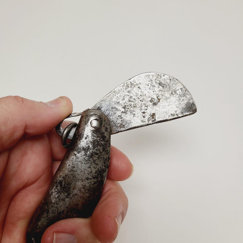 May include: An antique, weathered metal knife with a curved blade. The blade is open, revealing a textured surface. The handle is dark and appears worn. A small metal ring connects the blade to the handle. The knife is held against a white background.