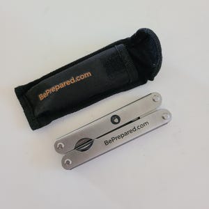 May include: A silver multi-tool with a black handle and the text "BePrepared.com" printed on it. The multi-tool is folded and comes with a black pouch.
