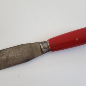 May include: A paint scraper with a rectangular metal blade and a red wooden handle. The handle has a hole at the end for hanging. The blade shows signs of use.