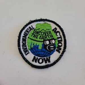May include: A white, black, and green embroidered patch with the text "Environmental Action Now" and a graphic of a globe with a gas mask over it. The globe is green and blue with the text "Discover the Earth" on it.