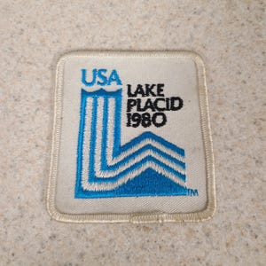 May include: A square, off-white patch with the text "USA LAKE PLACID 1980" in blue and black. The design includes a stylized blue mountain range and vertical lines. The patch has a stitched border.