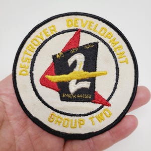May include: A round patch with the text "DESTROYER DEVELOPMENT GROUP TWO" in yellow. The patch features a black and red design with a yellow stripe and the number "2". The patch is on a cream-colored background with a black border.
