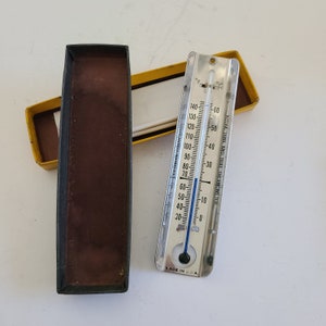 May include: A vintage metal thermometer with a blue liquid column, displaying Fahrenheit and Celsius scales. The thermometer is next to its original box, which is black and brown. The box is open, revealing the thermometer and a yellow box.