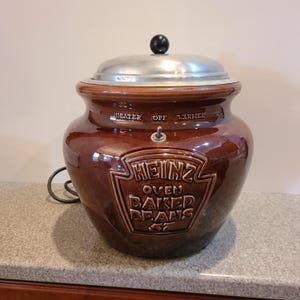 May include: A brown ceramic Heinz Oven Baked Beans crock pot with a silver lid and a black knob. The crock pot has a cord and a switch with the words "Heater", "Off", and "Warmer" printed on it.