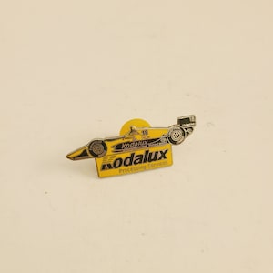 May include: A yellow and black pin featuring a race car with the text "Kodalux Processing Services" and the number "18" on the car.