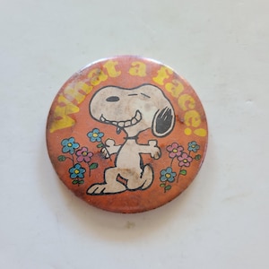 May include: A round orange button with a cartoon image of Snoopy with the text "What a face!" in yellow letters.