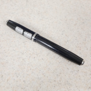 May include: A black fountain pen with silver accents. The pen has a shiny, smooth finish and a silver band near the center. Small white labels with text are visible on the pen's body.