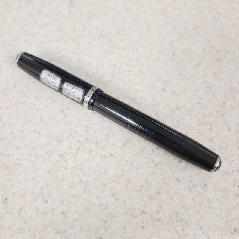 May include: A black fountain pen with silver accents. The pen has a shiny, smooth finish and a silver band near the center. Small white labels with text are visible on the pen's body.