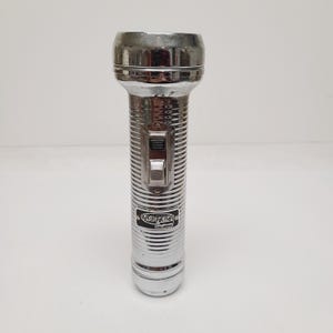 May include: A vintage-style silver-colored metal flashlight with a ribbed cylindrical body. It features a sliding power switch on the side and a black rectangular label near the base with the white text TRUTEST Supreme printed on it.