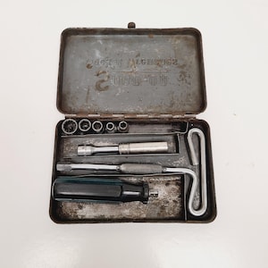 May include: An open, vintage metal toolbox with a hinged lid. Inside are various tools, including sockets, a ratchet, a screwdriver, and a T-handle wrench. The toolbox is gray with some rust and wear, suggesting age and use. The text on the lid is blurry.