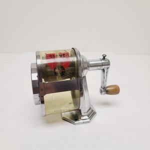 May include: Vintage Giant pencil sharpener with a clear plastic barrel and a silver metal base. The barrel has a red label with the word "GIANT" visible. The sharpener has a wooden handle and is marked "MADE IN U.S.A."