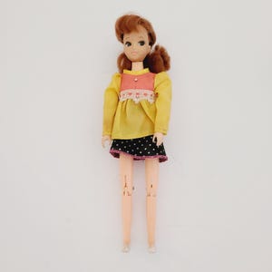 May include: A vintage plastic doll with brown hair, wearing a yellow and pink dress with a white lace trim, a black and white polka dot skirt, and pink shoes.