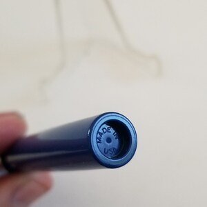 May include: Close-up of a navy blue pen with the text "MADE IN USA" on the end. The pen is cylindrical and appears to be made of plastic. A blurred object is in the background.