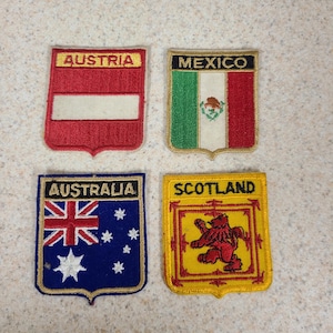 May include: Four embroidered patches featuring flags and country names. Austria's patch is red with a white stripe and the word "AUSTRIA". Mexico's patch has green, white, and red stripes with the word "MEXICO". Australia's patch is blue with the Union Jack and the word "AUSTRALIA". Scotland's patch is yellow with a red lion and the word "SCOTLAND".