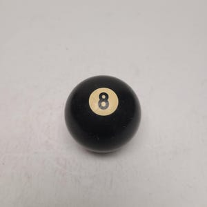 May include: A solid black billiard ball featuring a circular white inlay in the center. The white inlay contains the number 8 printed in a bold black font. The surface of the ball is smooth and glossy, reflecting light across its spherical shape.