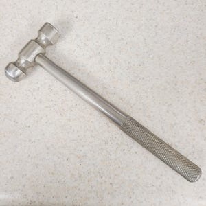 May include: A silver ball-peen hammer with a textured handle. The hammer has a rounded head on one side and a flat striking surface on the other. The handle is straight and tapers slightly towards the end.