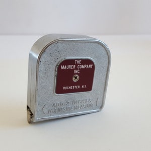 Puede incluir: Un flex&oacute;metro vintage de metal plateado con una etiqueta granate que dice "THE MAURER COMPANY INC. ROCHESTER, N.Y.". Tambi&eacute;n es visible el texto "ADD 2 INCHES FOR INSIDE MEASURE". Una herramienta cl&aacute;sica para medir.