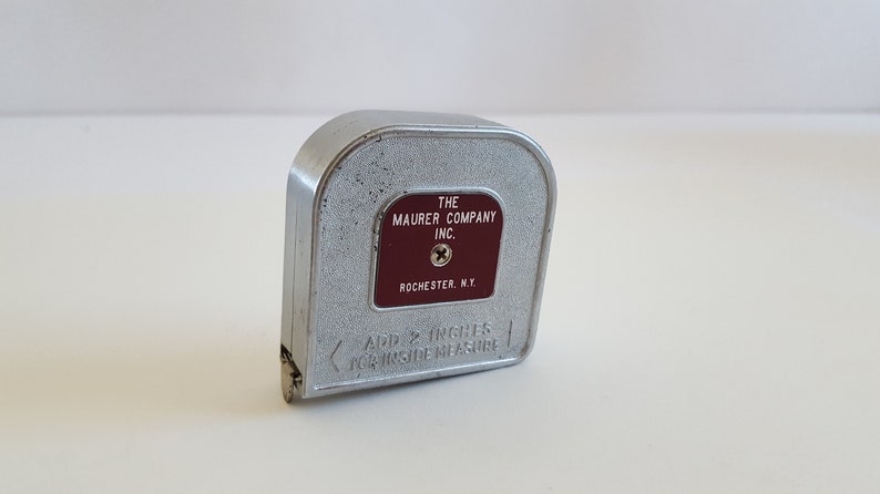 Puede incluir: Un flex&oacute;metro vintage de metal plateado con una etiqueta granate que dice "THE MAURER COMPANY INC. ROCHESTER, N.Y.". Tambi&eacute;n es visible el texto "ADD 2 INCHES FOR INSIDE MEASURE". Una herramienta cl&aacute;sica para medir.