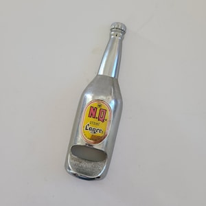 May include: A silver bottle opener shaped like a beer bottle. The opener has a yellow and red label that reads "N.Q. Lager 370ml". The bottle opener is made of metal and has a bottle cap on top.