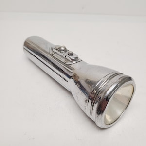 May include: A vintage, chrome-plated flashlight with a cylindrical body and a flared head. The flashlight has two buttons on the top and the text "MADE IN U.S.A." is visible. The flashlight is designed for handheld use.