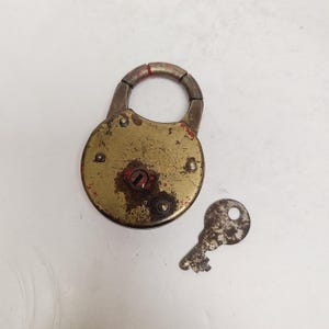 May include: An antique brass padlock with a matching key. The lock is round with a hinged shackle and shows signs of age, with a worn, tarnished finish and red paint accents. The key is small and also shows signs of wear.