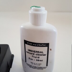 May include: A white bottle of Realistic Universal Record Cleaner Fluid, 2 oz (59 ml). The bottle has a green cap and black text.