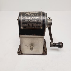 May include: Vintage black and silver pencil sharpener with the text "Pencil Sharpener" and "Automatic Pencil Sharpener Co. Chicago, U.S.A." The sharpener has a crank handle and a receptacle for shavings. The base has four screw holes.