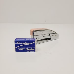May include: A silver Swingline stapler with a peach-colored top, next to a blue box of Cub Staples. The stapler has the words "Lift to Load" printed on it. The box of staples has the Swingline logo and the words "Cub Staples" printed on it.