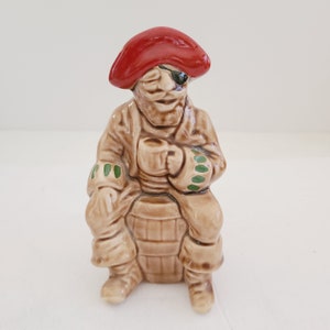 May include: A ceramic figurine of a pirate wearing a red hat and an eyepatch, sitting on a barrel and holding a cup.