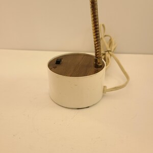 May include: A small, white desk lamp with a flexible, brass-colored neck. The lamp has a round base with a wooden top and a small switch. A cream-colored cord extends from the base.
