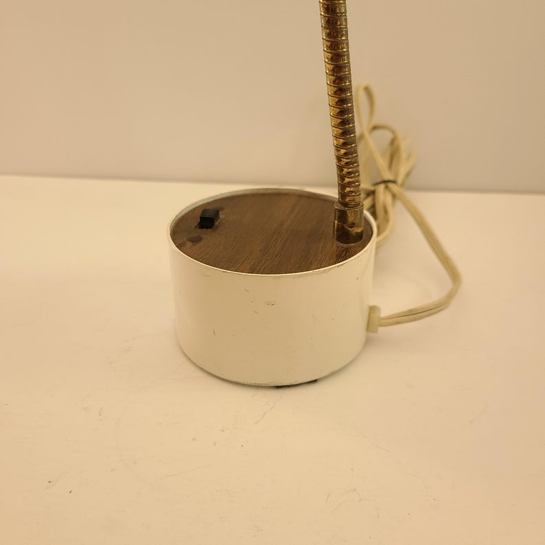 May include: A small, white desk lamp with a flexible, brass-colored neck. The lamp has a round base with a wooden top and a small switch. A cream-colored cord extends from the base.