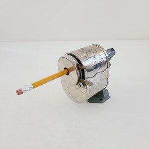Puede incluir: Un sacapuntas vintage de plata con un l&aacute;piz amarillo insertado. El sacapuntas tiene una manivela y una base de metal.