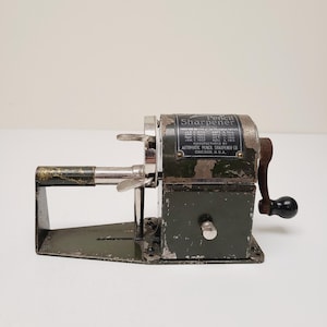 May include: Vintage green and silver pencil sharpener with a black crank and the text "Pencil Sharpener". The sharpener is manufactured by Automatic Pencil Sharpener Co. Chicago, U.S.A.