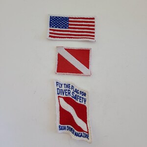 May include: Three embroidered patches. The top patch is a red, white, and blue American flag. The middle patch is a red and white scuba diver down flag. The bottom patch is a red and white scuba diver down flag with the text "Fly the flag for diver safety" and "Skin Diver Magazine" in blue.