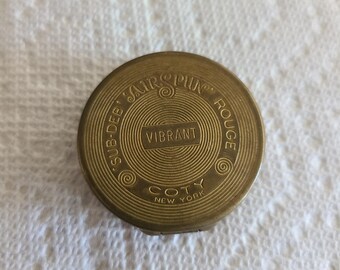 May include: A gold round compact with the words "Sub-Deb Air-Spun Rouge" and "Vibrant" in a circle around the word "Coty New York".