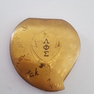 May include: A gold heart-shaped compact with the inscription "Sisterhood Forever" and the Greek letters "ΔΦΣ" in the center.