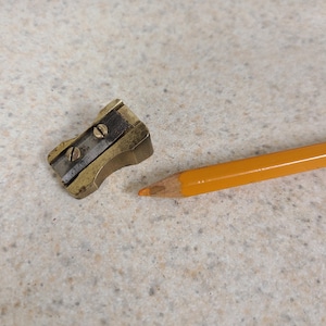 May include: A small, wedge-shaped brass pencil sharpener featuring a single steel blade secured by two flat-head screws, positioned next to the sharpened tip of a yellow wooden pencil with a visible orange lead core.