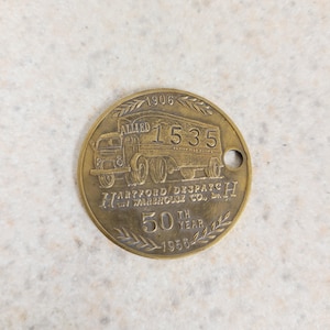 May include: A round, brass-colored token featuring a vintage truck design. The token includes the text "ALLIED 1535", "HARTFORD DISPATCH WAREHOUSE CO., Inc.", and "50TH YEAR 1956". A small hole is present near the edge.