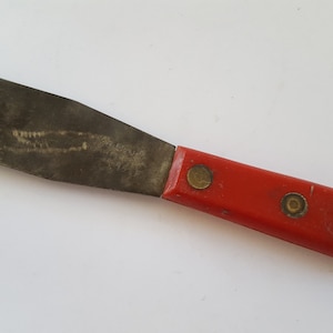 May include: A red-handled putty knife with a metal blade. The handle has three brass rivets.
