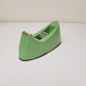 May include: A green tape dispenser with a clear plastic tape holder. The dispenser has a dark brown base and a metal cutting edge. The design is a simple, functional shape.