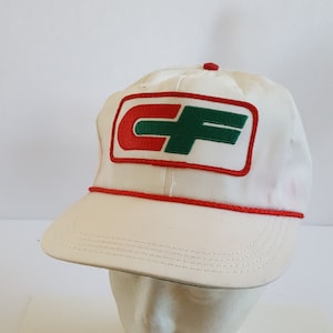 May include: A white baseball cap with a red and green logo patch. The cap has a red rope detail along the brim and a red button on top. The logo features the letters "CF" in red and green.