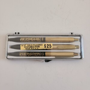 May include: A set of three gold-toned pens with black accents and a black and gold filigree design. The pens are in a clear plastic case with a white label that reads "14 KT. GOLD FILAGREE PERFUMED PENS MADE IN U.S.A." and a yellow price tag that reads "$25".
