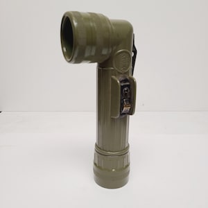 May include: An olive drab right-angle flashlight with a cylindrical body and a textured grip. The flashlight features a side-mounted black switch, a circular head with a ridged bezel, and a metal clip attached to the back for secure carrying.