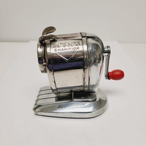 May include: A vintage silver-toned Boston Champion pencil sharpener. The sharpener has a red handle and the words "BOSTON CHAMPION" are printed on the front. The sharpener is made of metal and has a classic design.