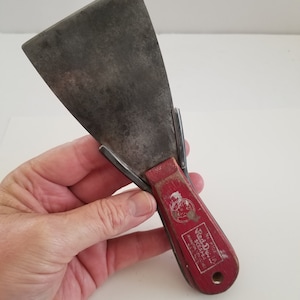 May include: A vintage Red Devil brand putty knife with a red wooden handle and a metal blade. The handle has the text "Red Devil Scraper" and "Rust-Resistant Steel" printed on it.