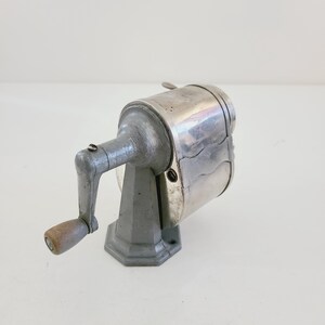 Puede incluir: Un sacapuntas vintage de plata con una manivela de madera. El sacapuntas tiene una base de metal y un cuerpo de metal plateado.