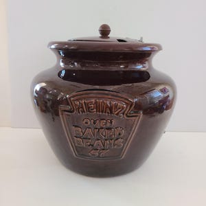 May include: A brown ceramic Heinz Oven Baked Beans 57 jar with a lid. The jar has a vintage look and is decorated with the Heinz logo and the words "Oven Baked Beans 57".