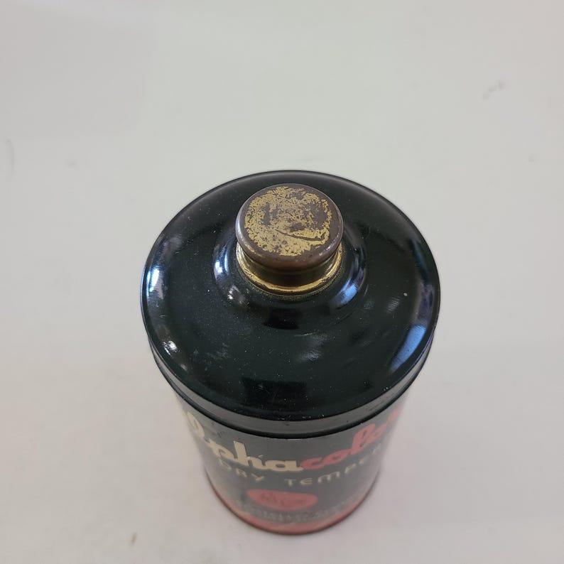 May include: A black metal tin with a gold lid. The tin has red and white text that reads "Alpha-Cola Dry Temper" and a red circle with a white star inside.