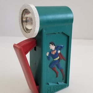 May include: A green plastic flashlight with a red handle and a Superman image on the side. The flashlight says "FLASHLITE" on the bottom.